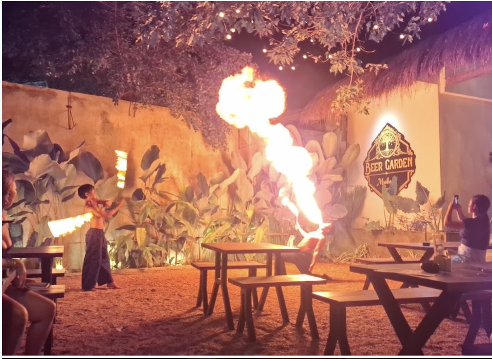 Fire dancer breathing a huge fireball in the Beer Garden at night, under string lights